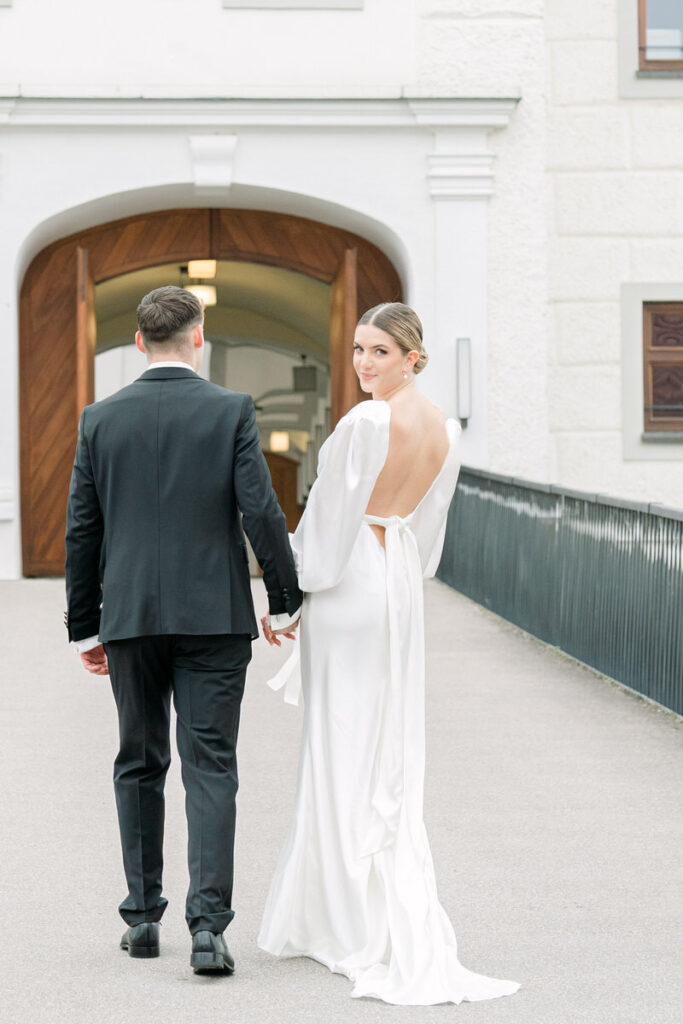 Bräutigam hält Hand der Braut während sie sich umdreht und zum Fotografen aus Rosenheim blickt mit einer Schlosstor im Hintergrund