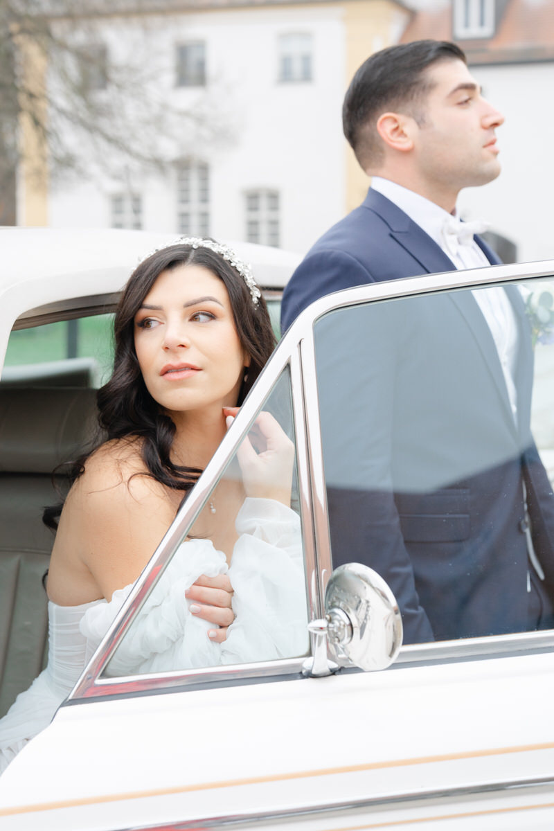 Braut sieht aus dem Fenster des Hochzeitsautos und lächelt während der Bräutigam im blauen Hochzeitsanzug geradeaus blickt