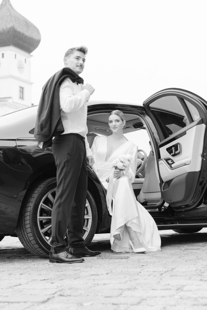 Braut sitzt in einer modernen Hochzeitslimousine und blickt in die Kamera vom Hochzeitsfotografen Rosenheim während der Bräutigam mit seinem Anzug posiert