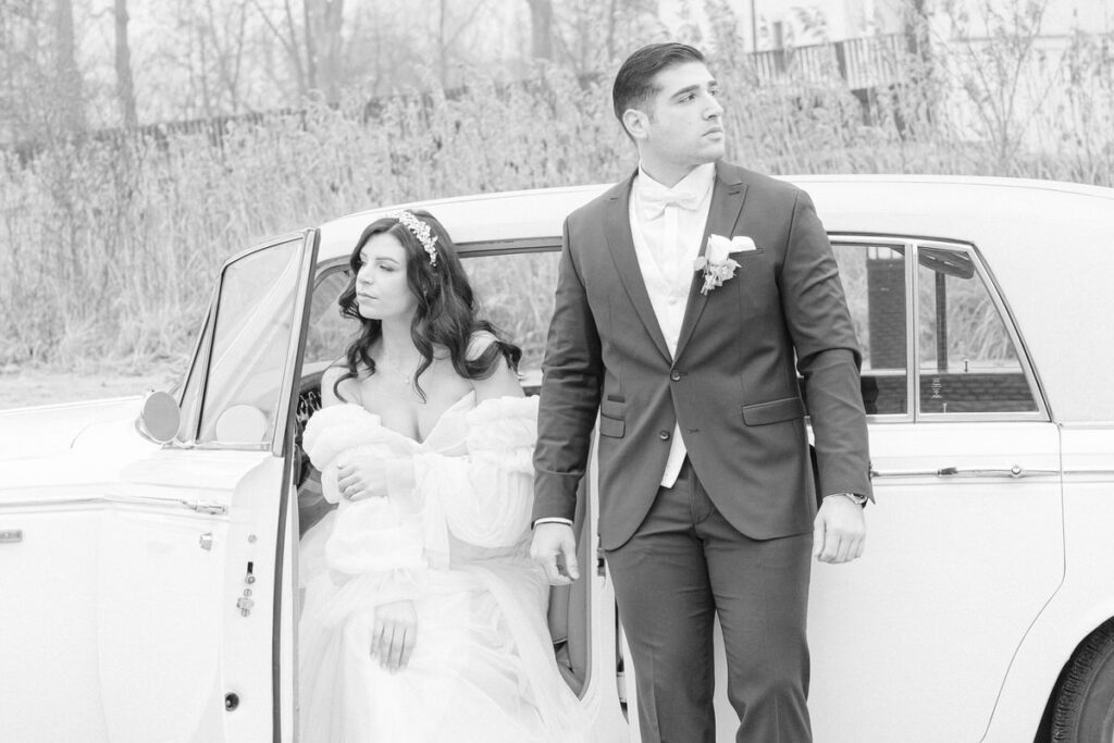 Eine Braut sitzt im Hochzeitsauto, während der Bräutigam sich an den Oldtimer lehnt und in die andere Richtung blickt, fotografiert in schwarz weiß
