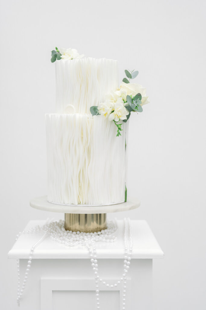 Hochzeitstorte auf einem Kuchenteller mit weißen eleganten Elementen, einer Perlenkette und weißer Blumendekoration bei einer Hochzeit im Schloss