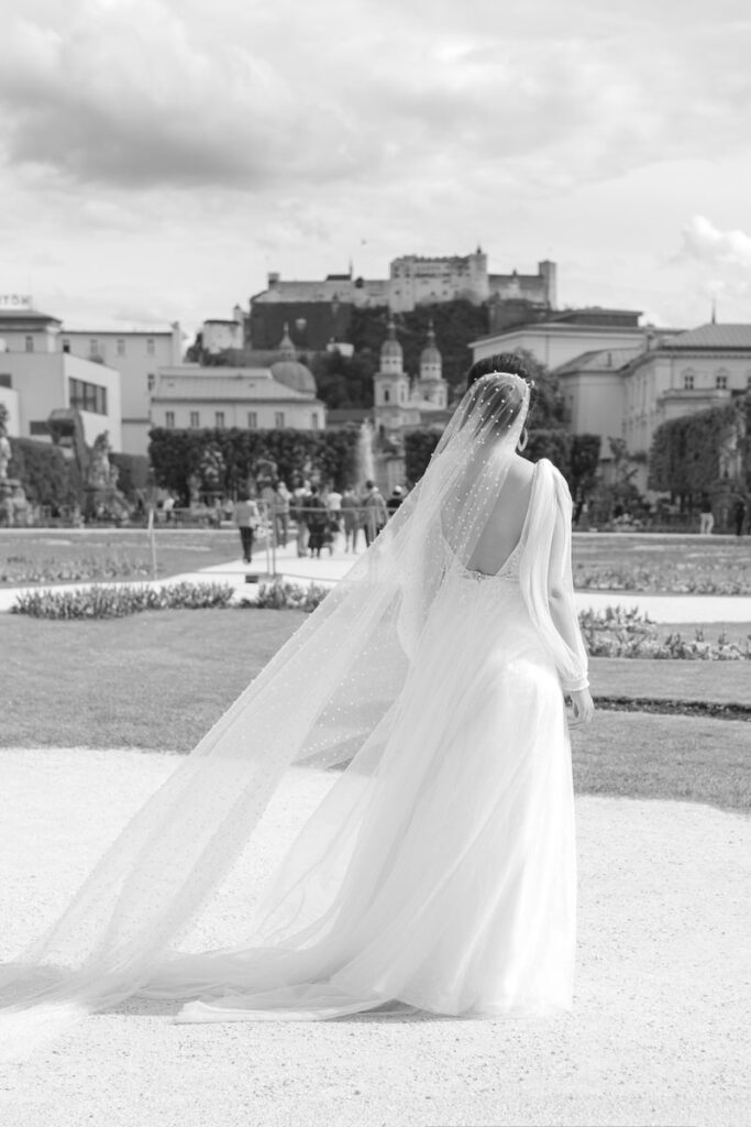 Rückansicht einer Braut mit einem Perlenschleier und einem eleganten Brautkleid mit Blick auf die Altstadt von Salzburg, fotografiert in schwarz weiß