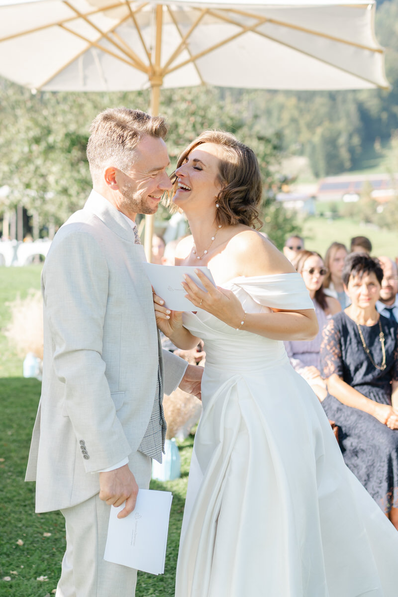 Hochzeitspaar lacht bei ihrem emotionalen Hochzeitsgelübte mit Hochzeitsgesellschaft im Hintergrund