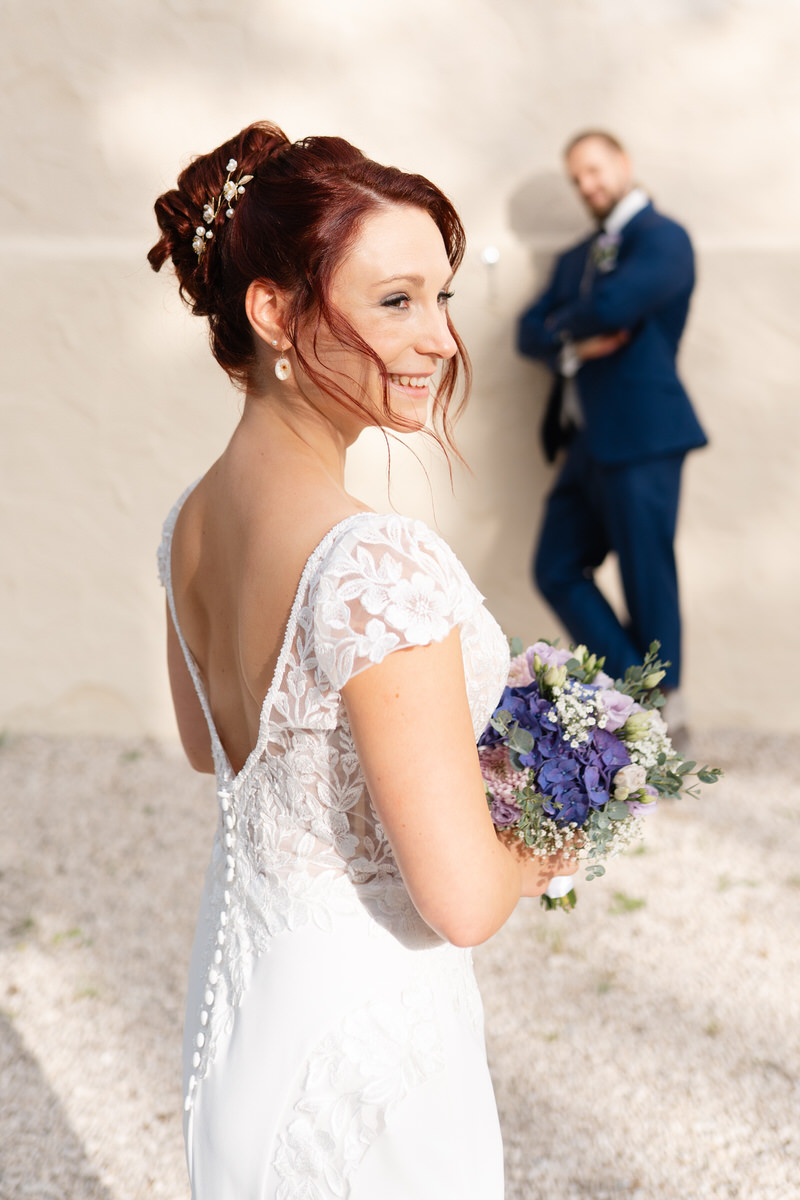 Braut hält Brautstrauß in den Händen und lächelt mit dem Bräutigam im Hintergrund