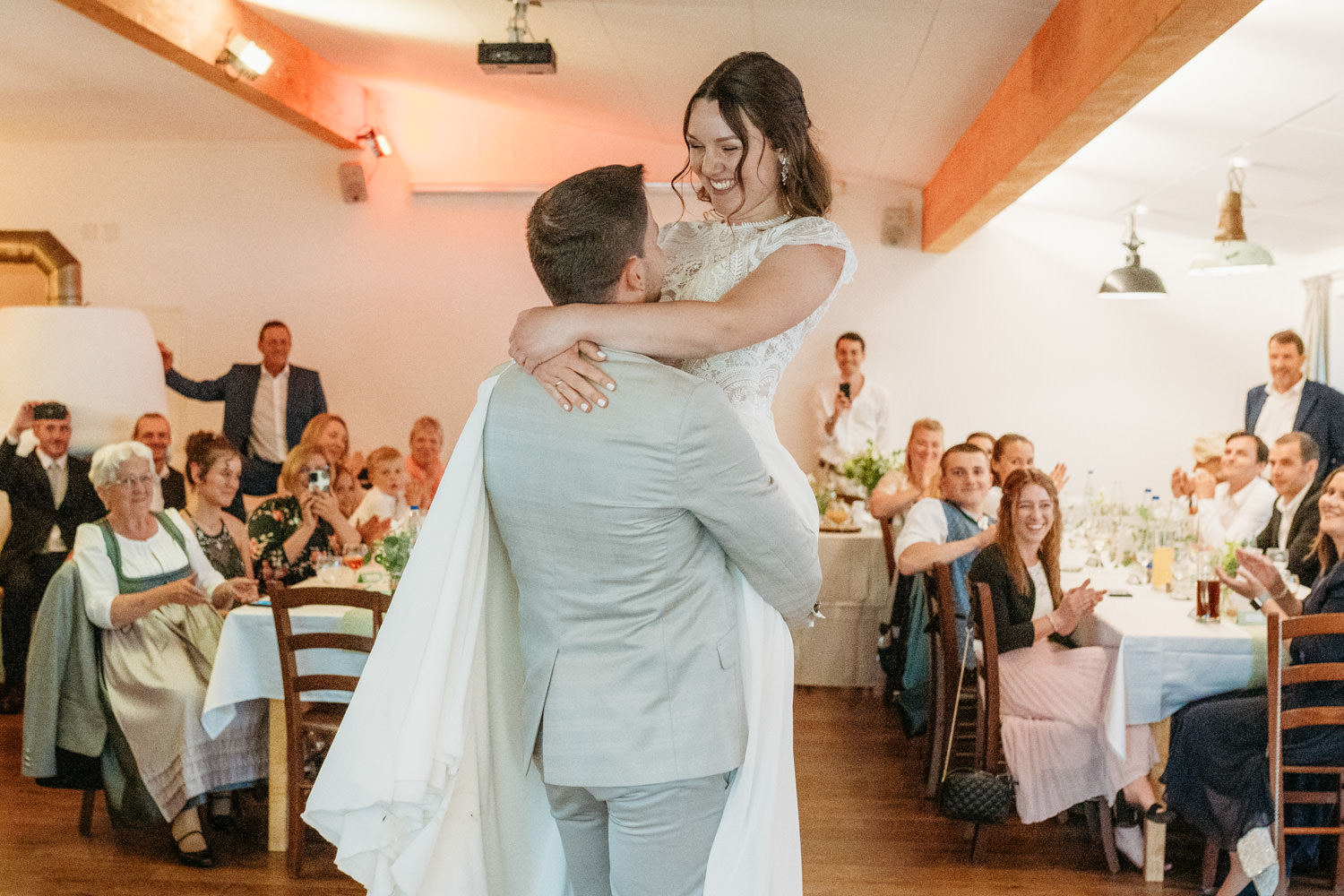 Bräutigam hebt die Braut beim Hochzeitstanz hoch, im Hintergrund sind Hochzeitsgäste